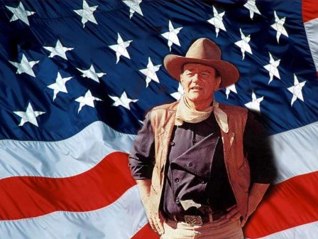 This is a classic image of John Wayne, in full Western uniform, standing in front of a giant flag of the United States of America.